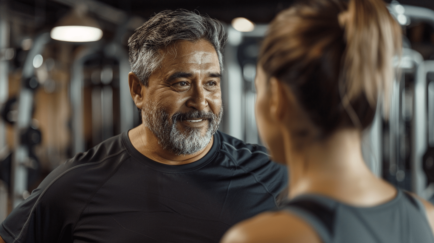 Hispanic man inside the gym talking with a fitness trainer.